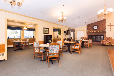 A spacious dining room with several round tables and wooden chairs with floral upholstery. The room features a brick fireplace with a clock above it and a cross on the wall to the right. There are chandeliers hanging from the ceiling and large windows with blue valances allowing natural light to enter. The walls are painted a light yellow with wood paneling along the lower half.