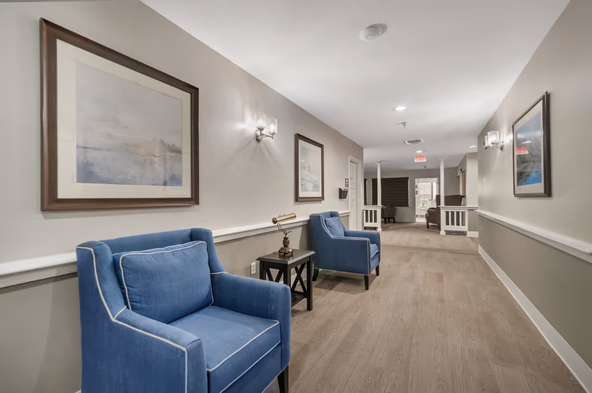 A hallway in a senior living facility with two blue armchairs and a small black side table with a decorative lamp between them. The walls are painted in neutral tones with framed artwork and wall sconces providing lighting. The floor is wood, and the hallway leads to a more open area with additional seating.