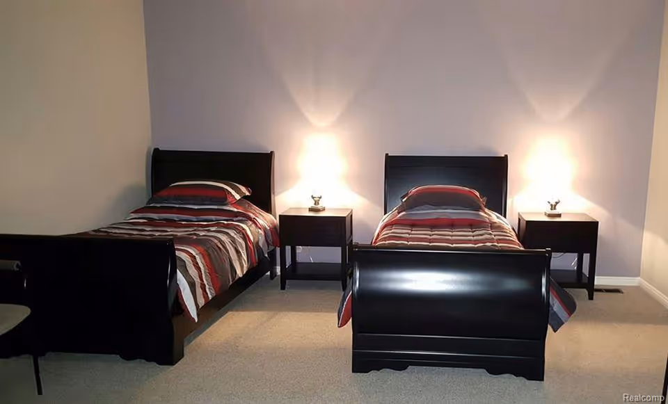A bedroom with two single beds, each with a striped comforter in shades of red, gray, and white. Between the beds is a small dark wooden nightstand with a lit lamp on top. The walls are light-colored, and the floor is carpeted in a neutral tone.