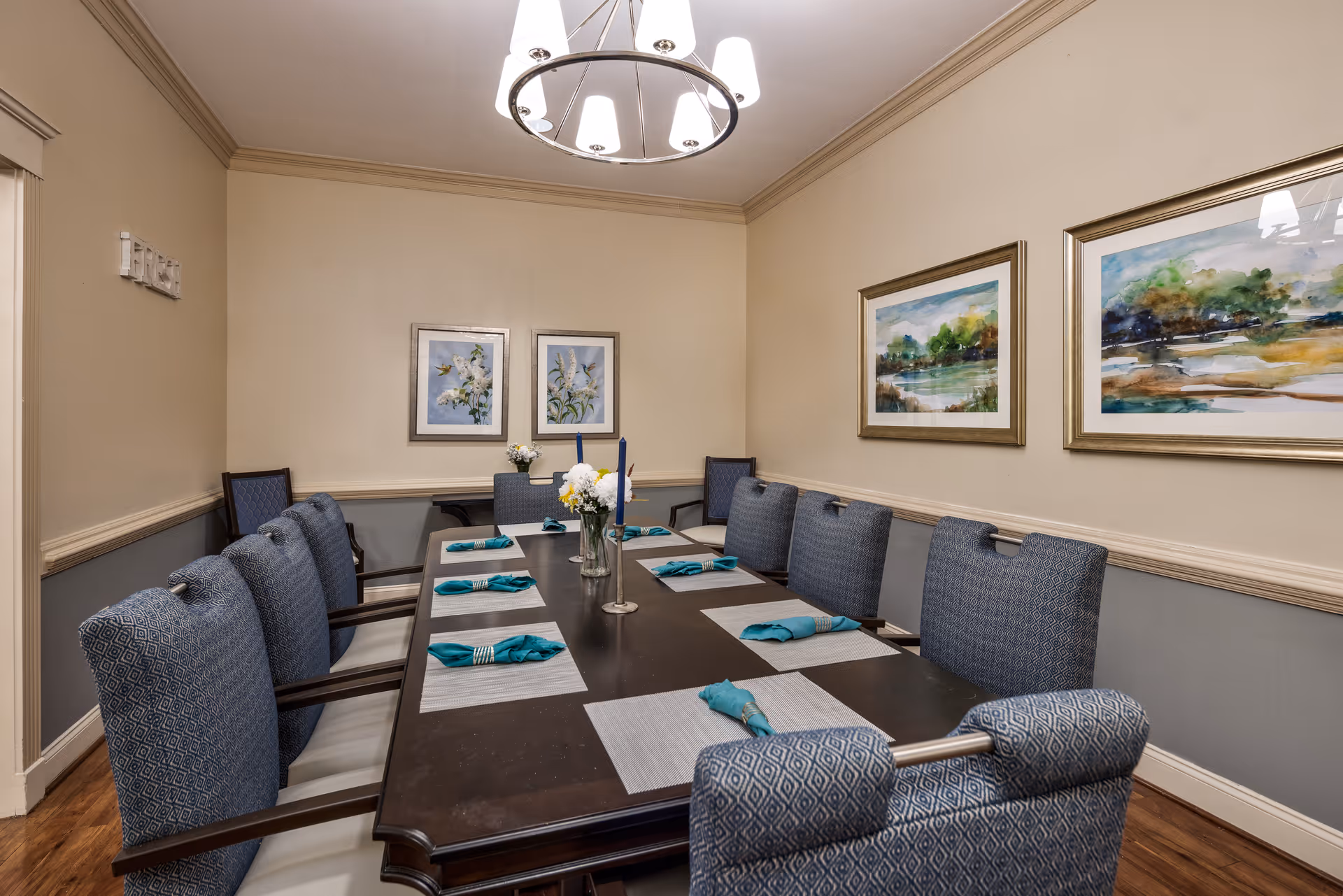 A formal dining room with a long dark wooden table set with placemats and teal napkins. The table is surrounded by cushioned chairs with patterned upholstery. The walls are beige with a chair rail and decorated with framed floral and landscape paintings. A modern chandelier hangs above the table.