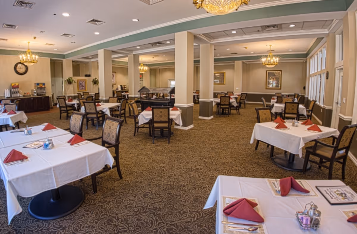 A spacious dining room with multiple tables covered in white tablecloths and set with red folded napkins, silverware, and menus. The room features patterned carpet, beige walls with white trim, chandeliers hanging from the ceiling, and large windows on one side. There is a buffet area with beverage dispensers and a clock on the wall in the background.