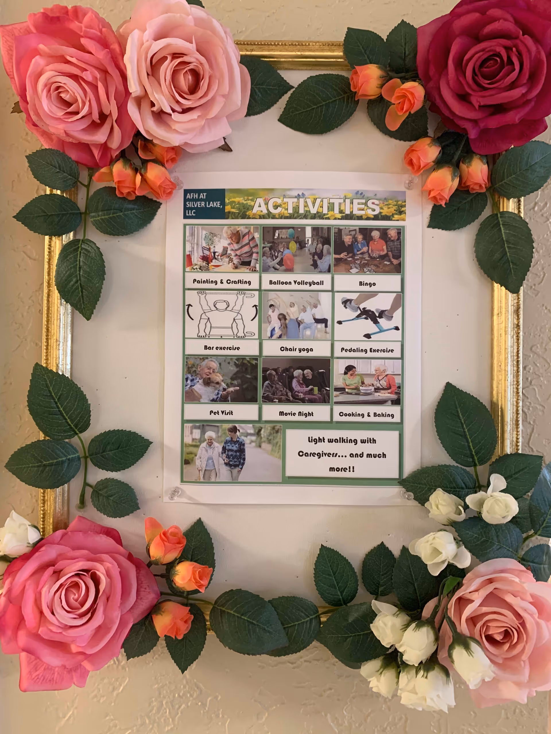 A decorative gold frame adorned with artificial pink, red, orange, and white roses and green leaves surrounds a printed activities schedule for AFH at Silver Lake, LLC. The schedule lists activities such as Painting & Crafting, Balloon Volleyball, Bingo, Bar exercise, Chair yoga, Pedaling Exercise, Pet Visit, Movie Night, Cooking & Baking, and Light walking with Caregivers. Each activity is accompanied by a small photo or illustration.