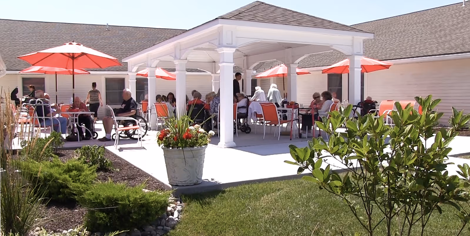Seniors sitting and socializing under a white pavilion with red umbrellas on an outdoor patio.