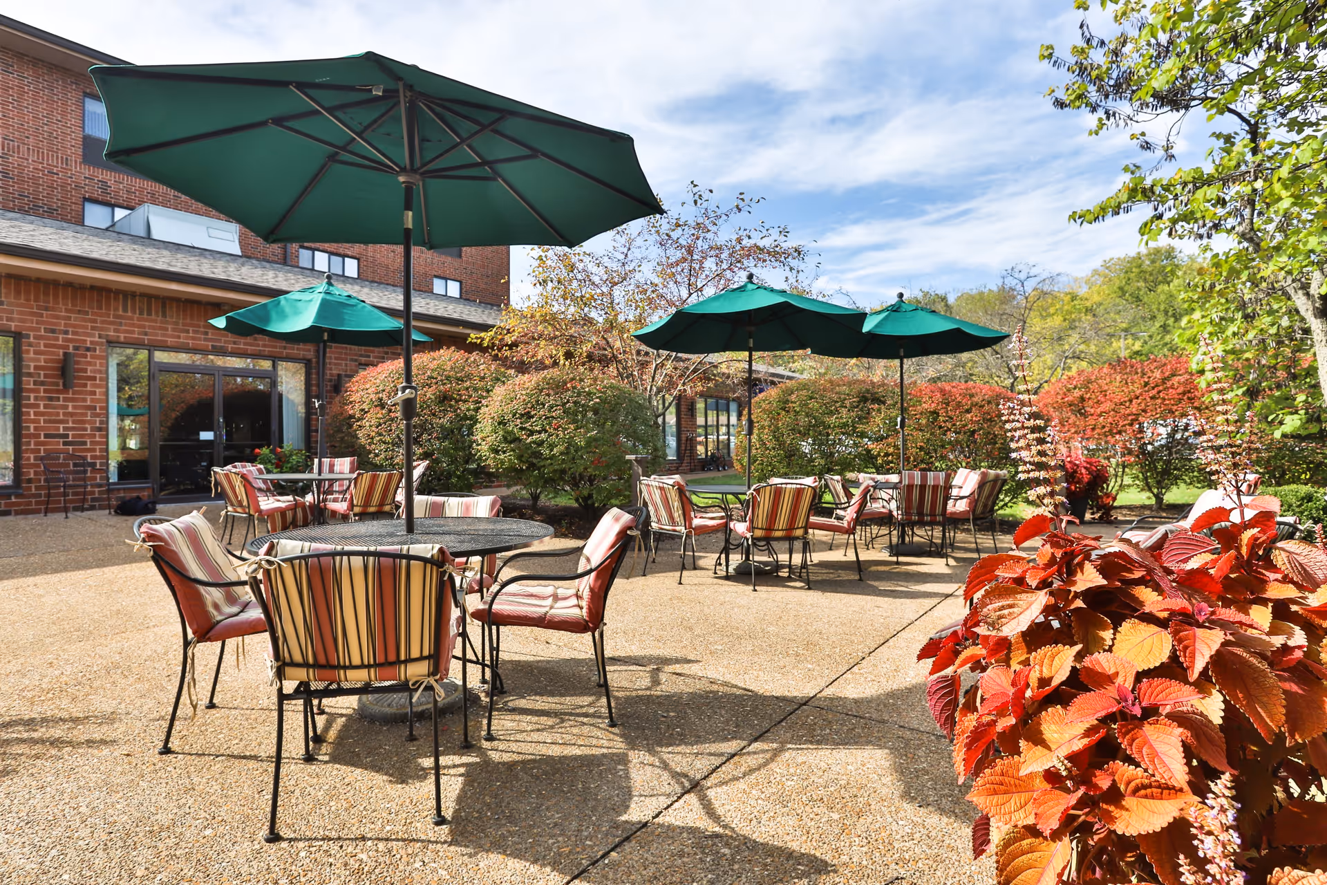 Outdoor patio area at Our Lady of Life Apartments with several round tables and striped cushioned chairs under large green umbrellas. The patio is surrounded by bushes and trees with autumn foliage, and a brick building is visible in the background under a partly cloudy sky.