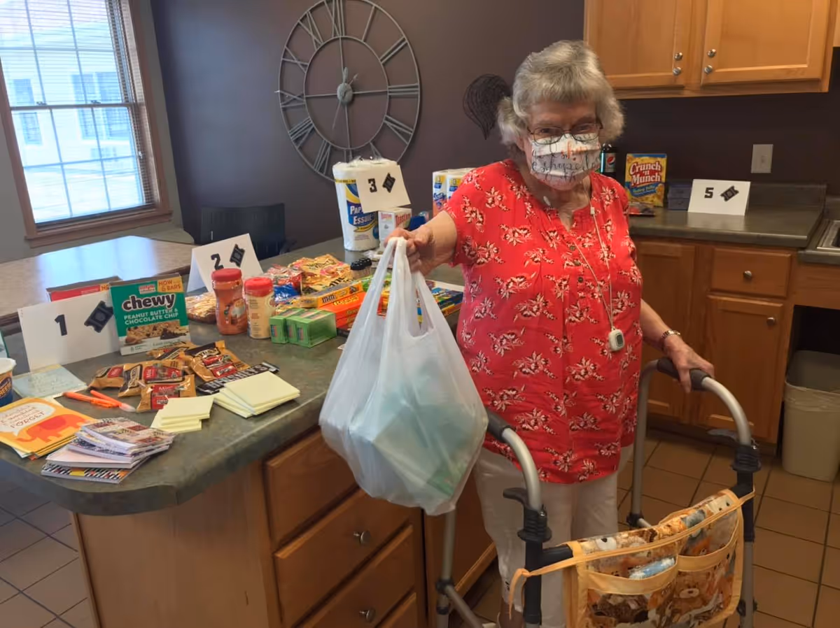 An elderly woman wearing a red floral shirt and a face mask stands in a kitchen area holding a plastic bag in one hand and using a walker with the other. On the counter beside her are various food items and household supplies organized with numbered cards.