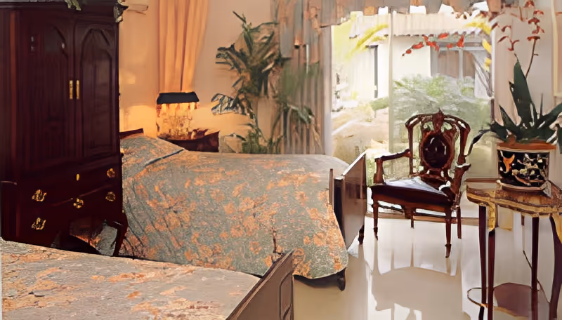 A bedroom with two beds covered in floral bedspreads, a dark wooden armoire, a vintage wooden chair, a small table with a potted plant, and large windows letting in natural light with greenery visible outside.