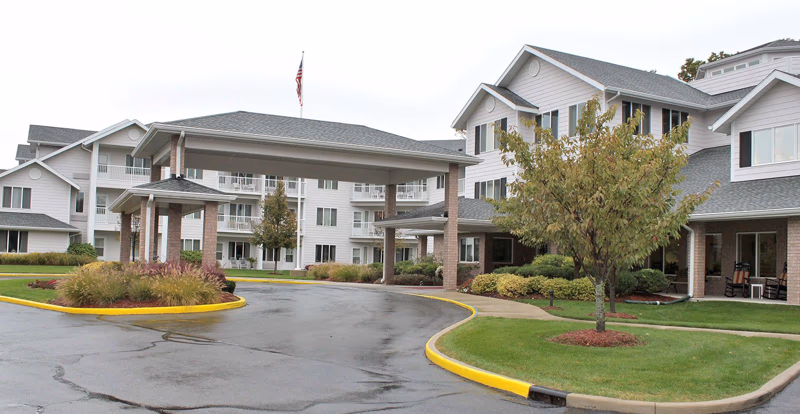 Exterior view of Holiday Alexis Gardens senior living facility showing a large covered entrance driveway, manicured lawns, trees, and a multi-story building with balconies and windows under an overcast sky.