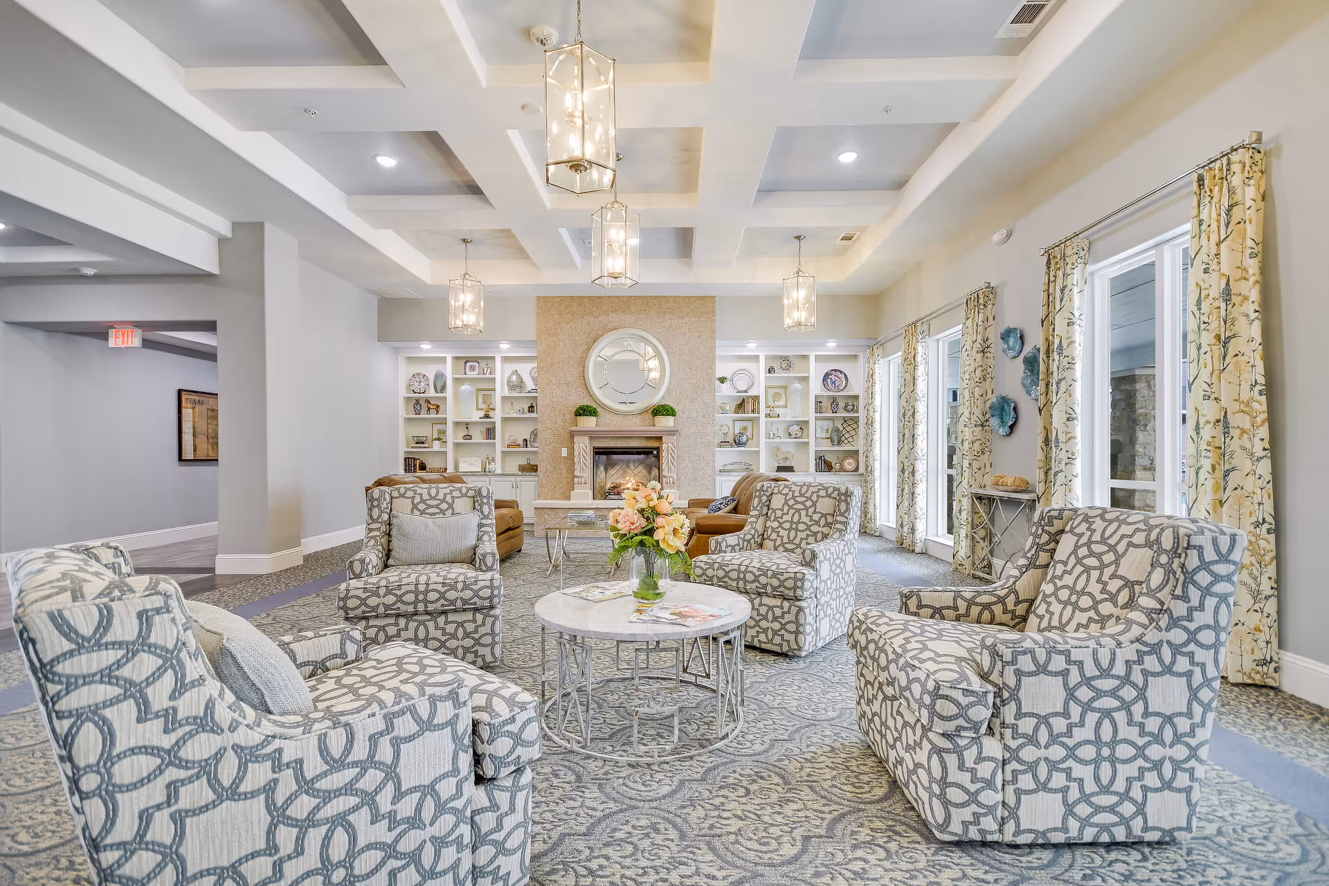 A bright and spacious senior living facility common area with patterned armchairs arranged around a round marble coffee table with a flower vase. The room features a fireplace with a large round mirror above it, built-in shelves with decorative items, large windows with floral curtains, and modern pendant lights hanging from a coffered ceiling.