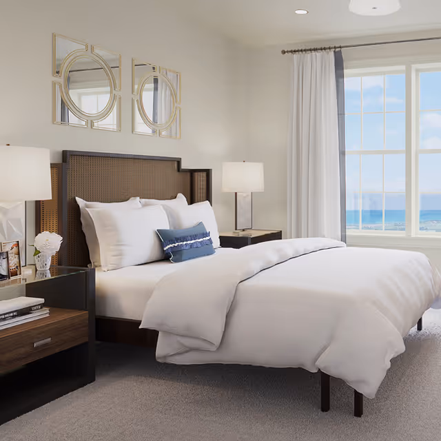 A bright and neatly made bed with white linens and a blue decorative pillow in a bedroom. The bed has a dark wooden headboard with a woven design. There are two bedside tables with modern lamps on each side. A large window with white curtains offers a view of the ocean and blue sky outside. Two decorative mirrors with gold frames hang above the bed.