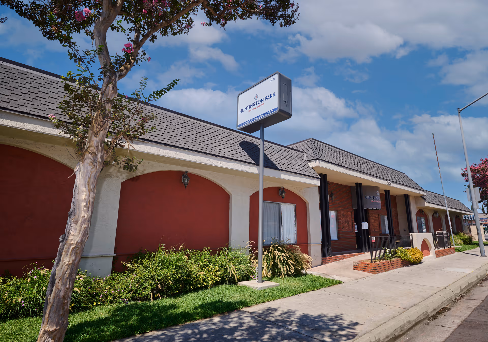 Street-level view of the Huntington Park Nursing Center building with a freestanding sign, landscaped front, and main entrance under a blue sky.