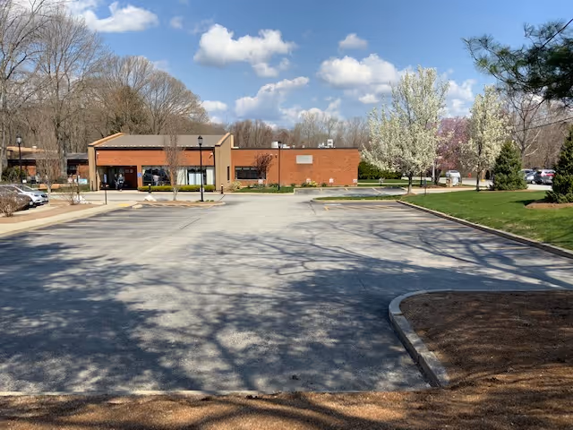 A parking lot in front of a single-story brick building with a sloped roof, surrounded by trees and a grassy area with blooming white and pink flowering trees under a partly cloudy blue sky.