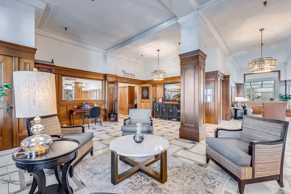 Bright, elegant senior living common area with armchairs around a round coffee table, wood-paneled walls, and chandeliers.