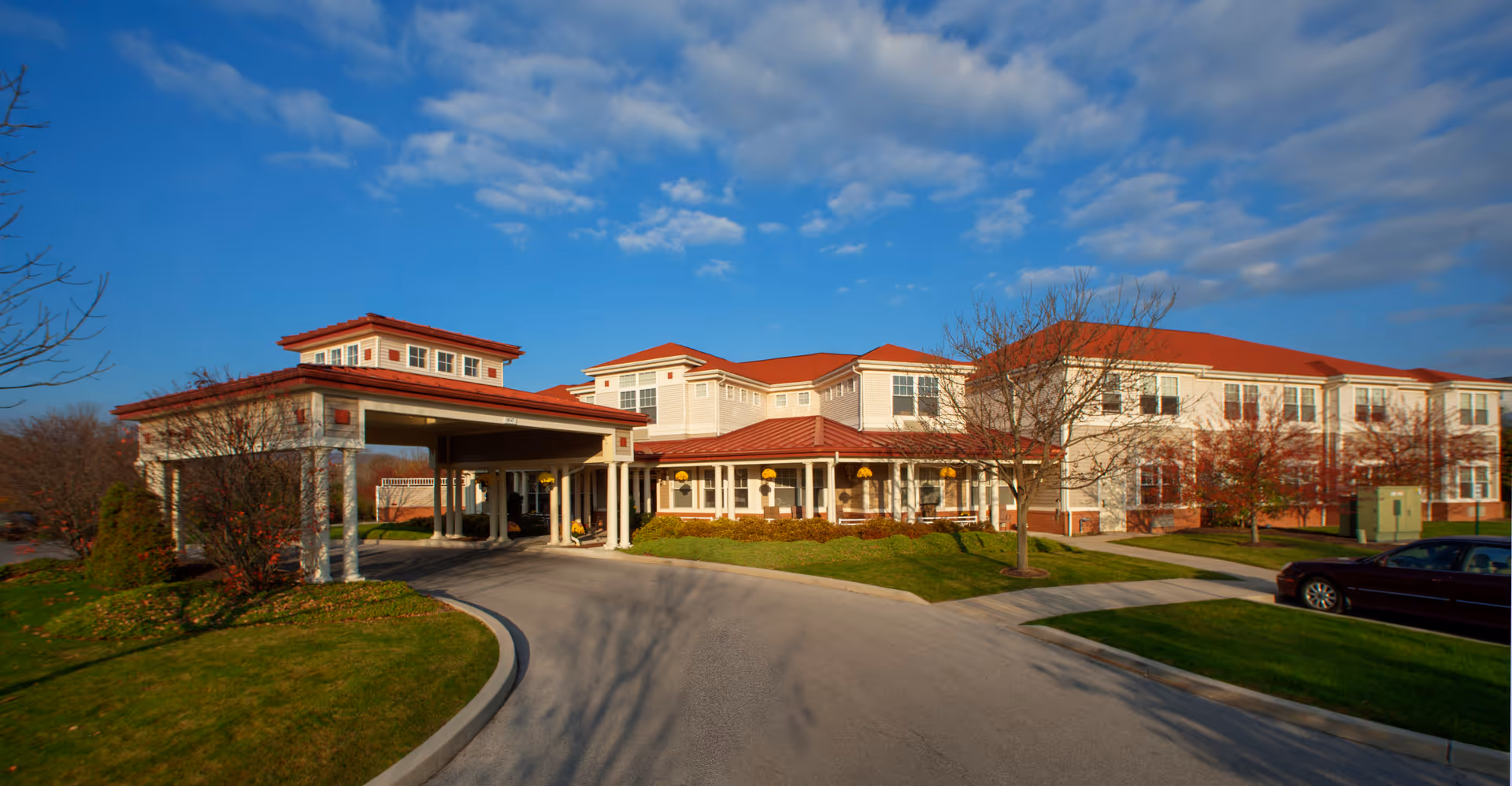 Front entrance of a large two-story senior living building with a covered porte-cochere, red roofs, landscaped lawns and a curved driveway under a blue sky.