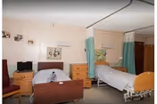 A room with two hospital-style beds separated by a curtain. Each bed has wooden headboards and footboards. There are two wooden dressers between the beds, a small TV on one dresser, and some pictures hanging on the wall. The room has a clinical, healthcare facility appearance.