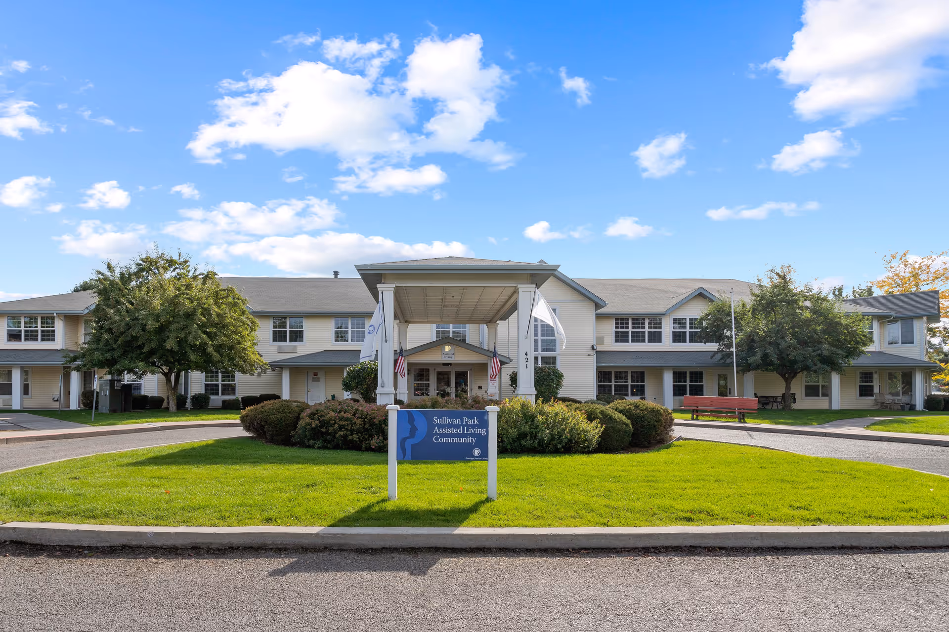 Front exterior view of Sullivan Park Senior Living Community building with a covered entrance, green lawn, bushes, trees, and a clear blue sky with some clouds.