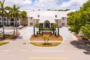 Front exterior view of Veranda Club facility with a white building featuring a large arched entrance, palm trees, landscaped garden beds, and a clear blue sky with some clouds.