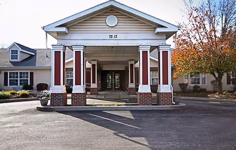 Front exterior view of The Wellington at Southport facility showing a covered entrance with white columns accented by red panels and brick bases, a paved driveway, and surrounding landscaping with trees and shrubs.