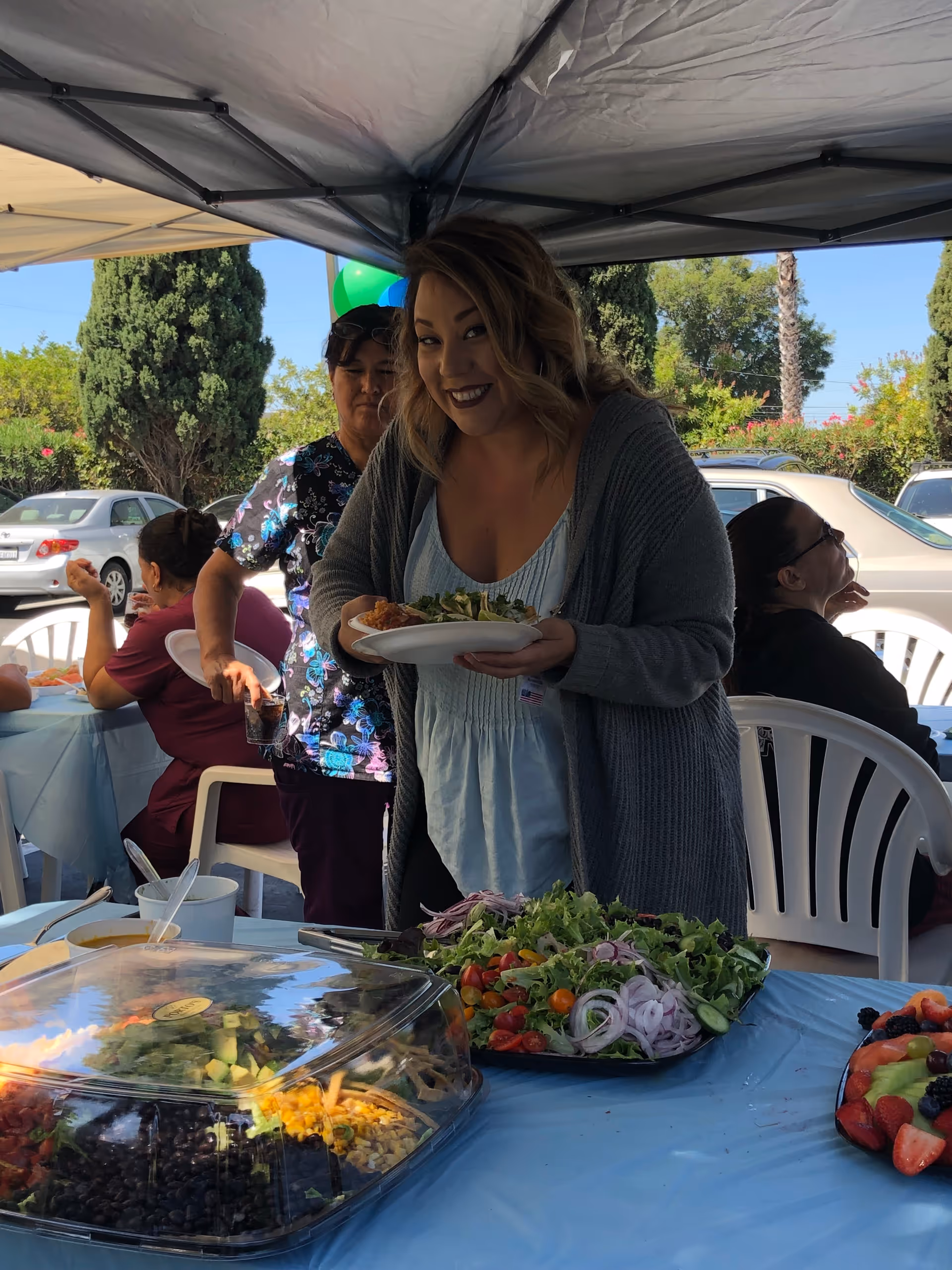 A woman smiling and holding a plate of food under a canopy tent at an outdoor event. There are other people seated at tables in the background, and a table in the foreground with a large salad platter and a covered dish of food. Trees and parked cars are visible in the background.