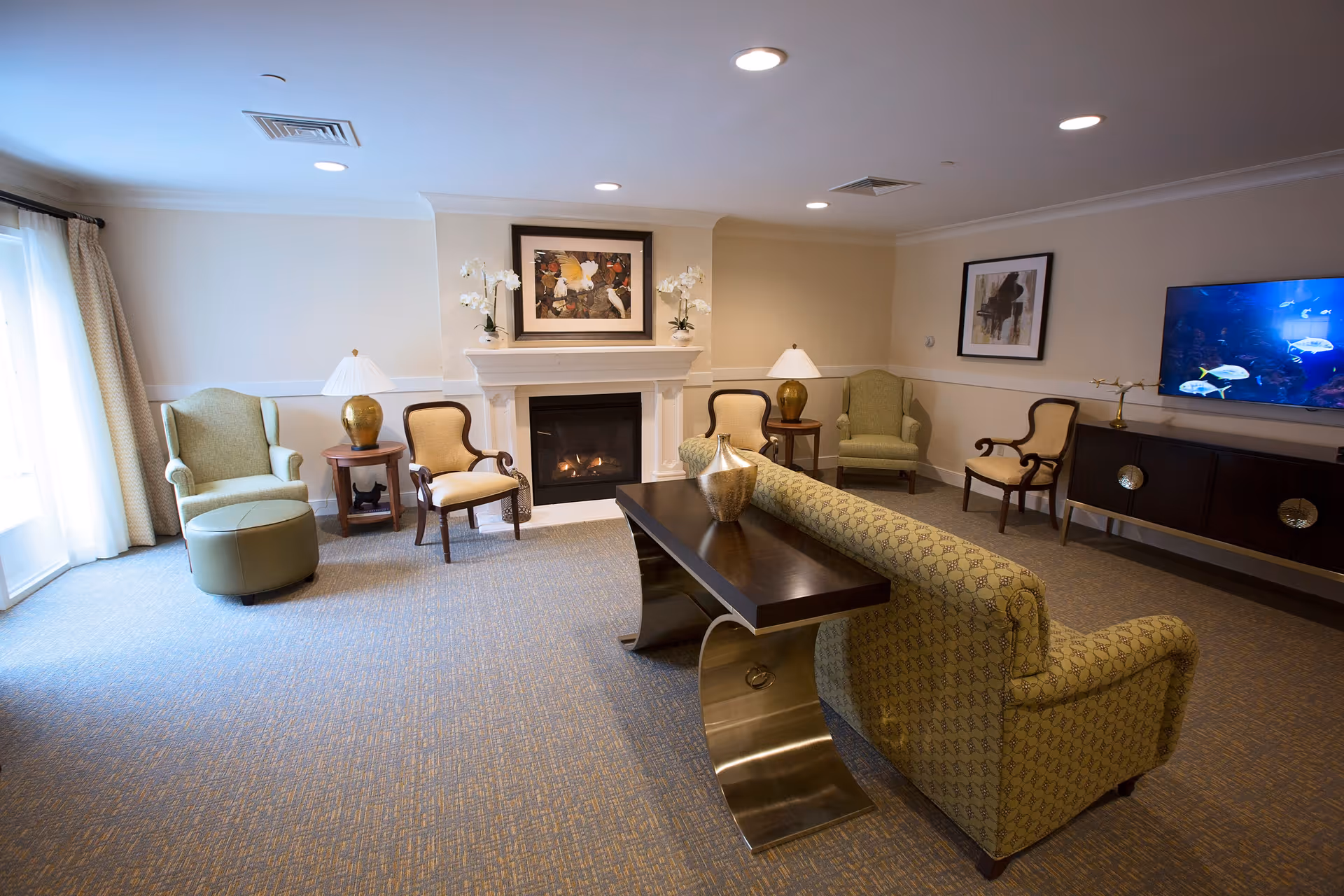 A cozy senior living common area with a patterned sofa facing a fireplace. There are two green armchairs and two wooden chairs with cream cushions arranged around the room. A dark wood console table with a decorative vase is placed behind the sofa. The room has beige walls, carpeted floor, framed artwork above the fireplace and on the wall, and a flat-screen TV displaying an underwater scene. Two table lamps with white shades are on side tables next to the chairs.