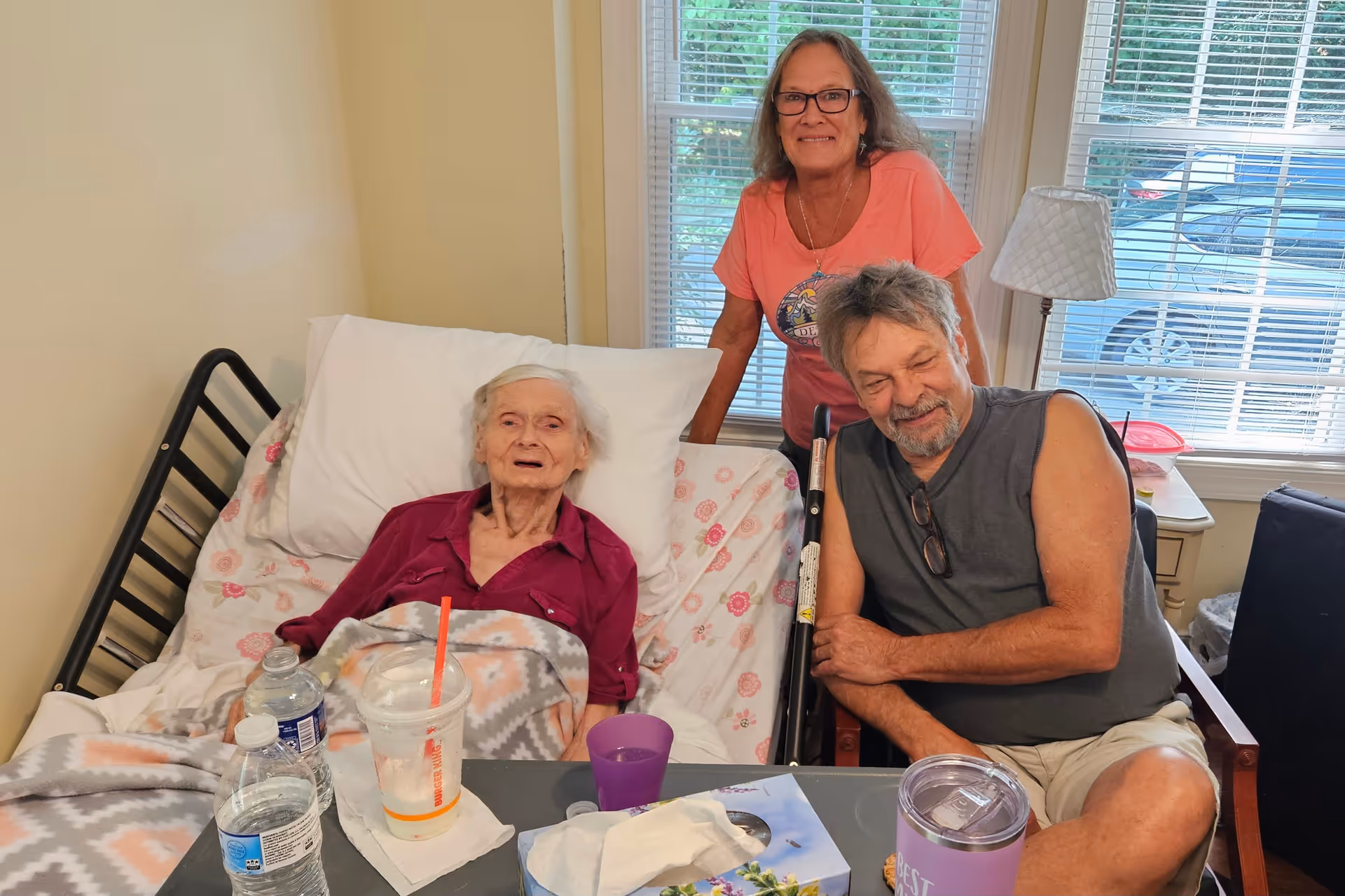 An elderly woman lying in a hospital-style bed covered with a patterned blanket, accompanied by a man sitting beside her and a woman standing behind the bed, all smiling. The room has a window with blinds, a lamp on a side table, and various drink containers on a table in front of the bed.