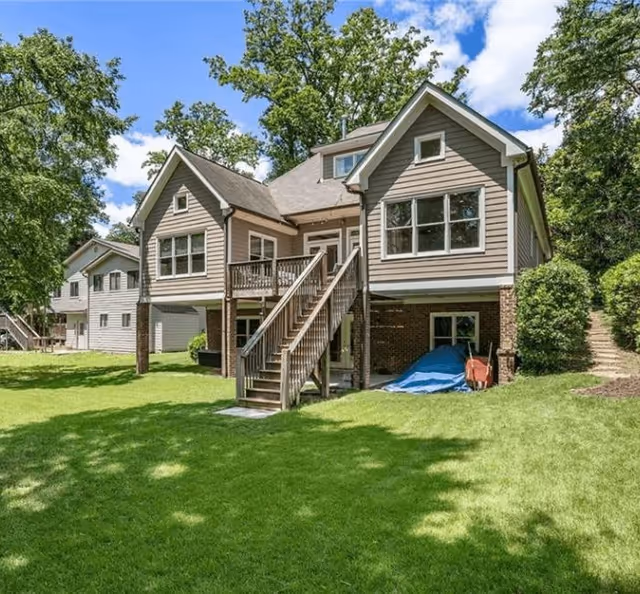 Two-story house exterior with wooden stairs up to a raised deck, green lawn in the foreground and trees under a blue sky.