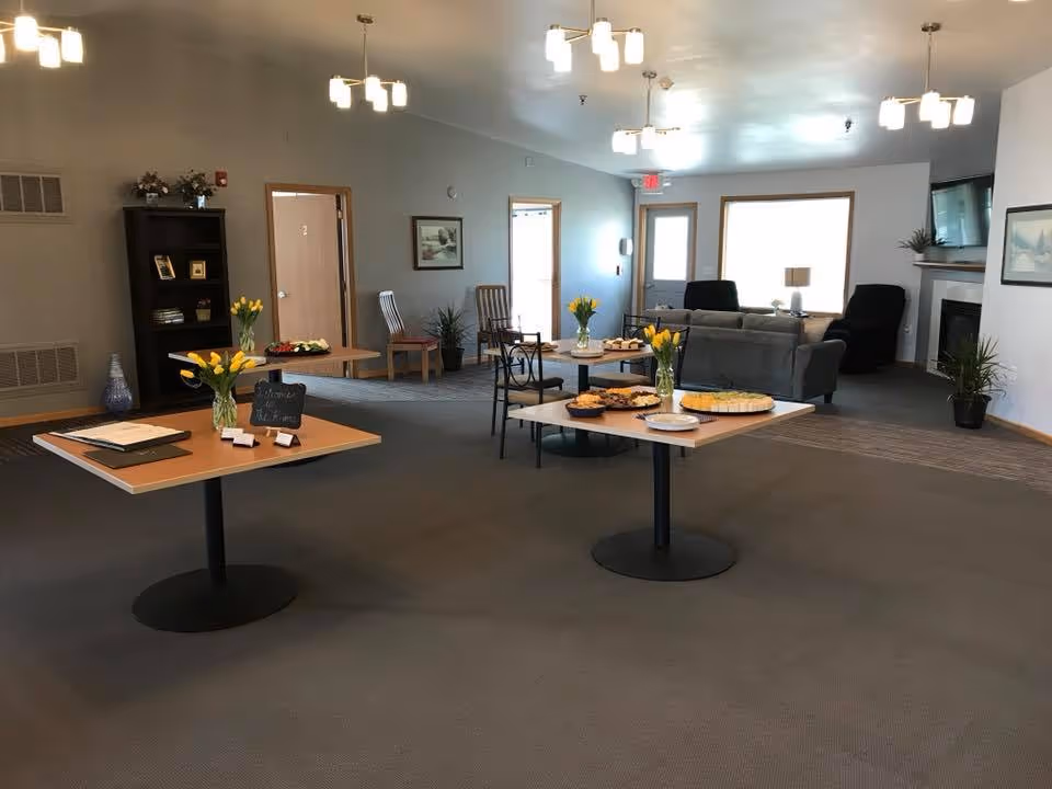 A spacious common area in a senior living facility with two tables set up with snacks and yellow flower centerpieces. The room has several chairs, a couch, a fireplace with a TV mounted above it, and large windows letting in natural light. The walls are decorated with framed pictures and there are potted plants placed around the room.