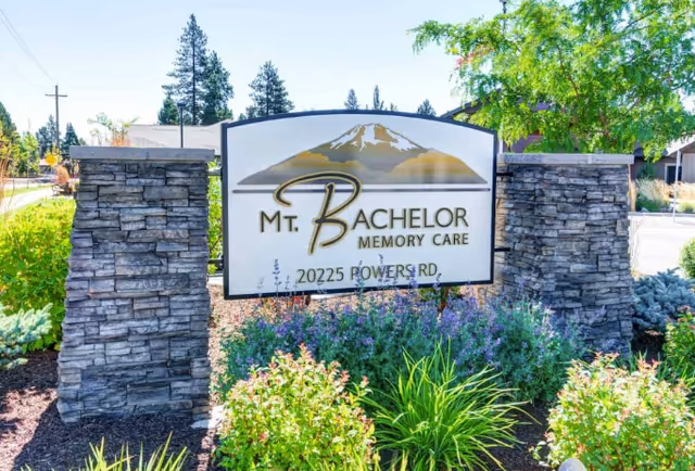 Outdoor sign for Mt. Bachelor Memory Care located at 20225 Powers Rd, surrounded by stone pillars and landscaped greenery with trees and plants in the background.