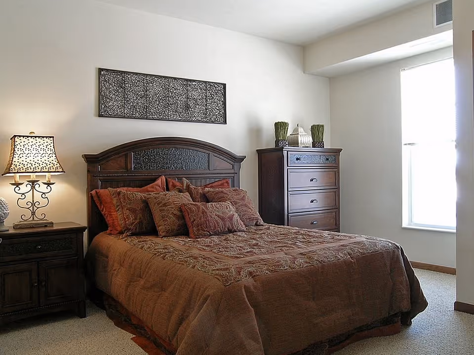 A bedroom with a large bed covered in a brown and patterned bedspread with multiple pillows. There is a wooden nightstand with a decorative lamp on the left side of the bed and a tall wooden dresser with decorative items on top against the wall. A window with blinds is on the right side, allowing natural light into the room. A piece of abstract wall art hangs above the bed.