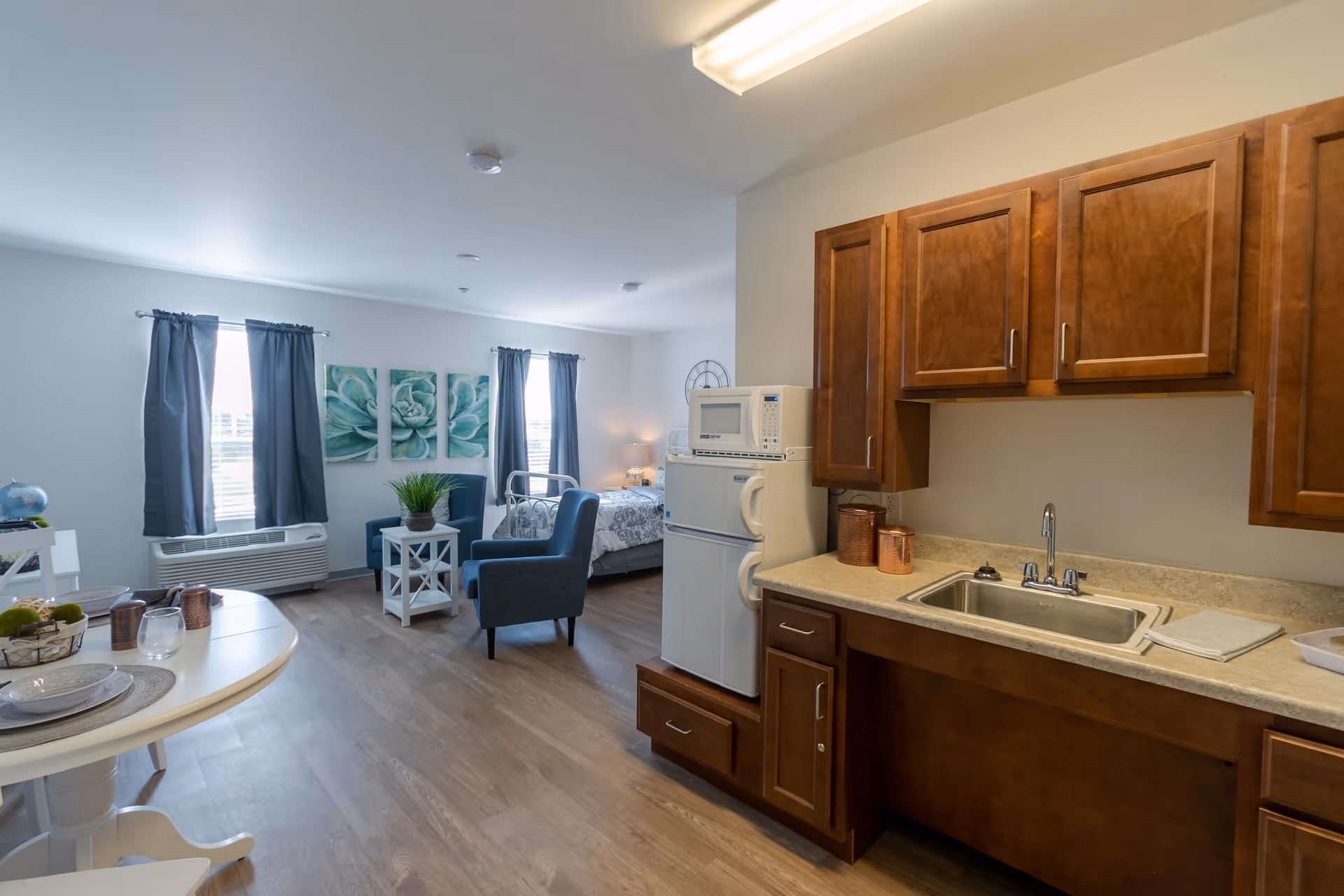A bright and clean studio apartment featuring a small kitchen area with wooden cabinets, a sink, a microwave, and a refrigerator. The living space includes two blue armchairs, a small white side table with a plant, and a bed near two windows with dark curtains. The floor is wooden, and the walls are painted white with decorative artwork above the chairs.