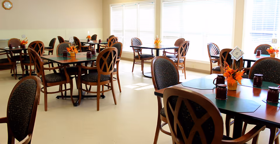 A bright dining room with multiple wooden tables and chairs arranged neatly. Each table has a small floral centerpiece with autumn-colored flowers and placemats. Large windows with blinds allow natural light to fill the room.