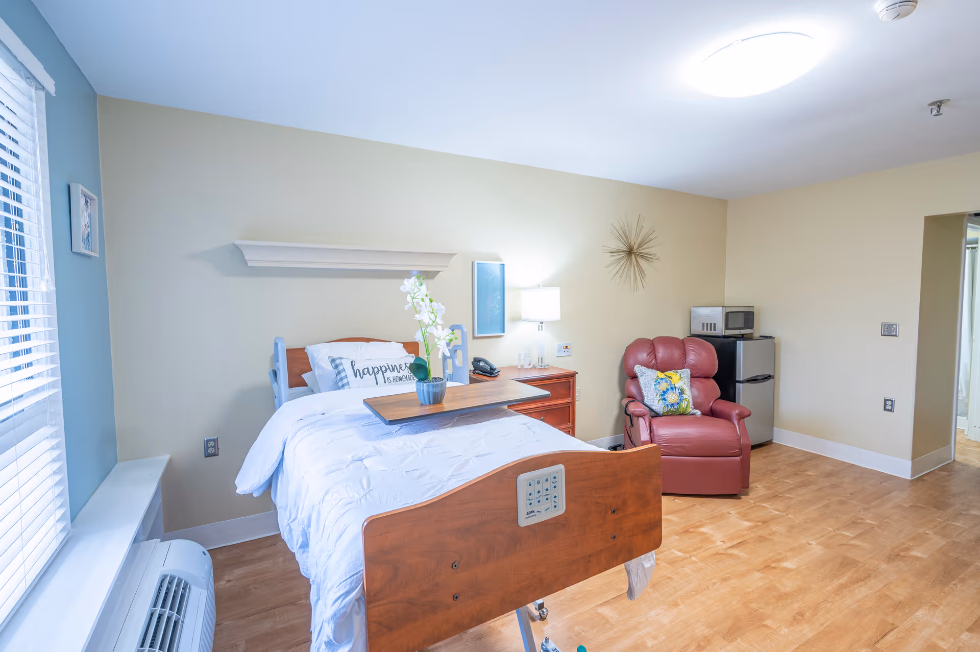 A bright and clean senior living bedroom featuring a hospital-style bed with white bedding and a decorative pillow that reads 'happiness is homemade'. Next to the bed is a wooden nightstand with a lamp and a phone. A red recliner chair with a colorful pillow is positioned near a small refrigerator and microwave. The room has light-colored walls, wood flooring, a window with blinds, and a decorative wall piece above the chair.