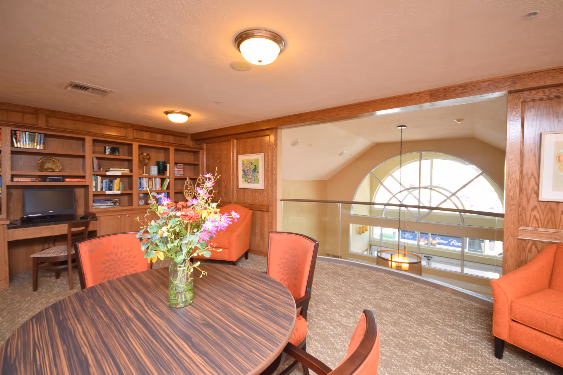 Interior view of a cozy common area in Stone River Retirement Community featuring a round wooden table with a vase of colorful flowers, four orange cushioned chairs, built-in wooden bookshelves with books and a small TV, and an orange armchair. The area overlooks a large arched window with a hanging light fixture and a view of the lower level.