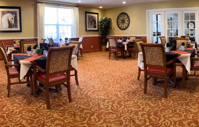 A dining room with several tables covered with white and black tablecloths, each set with chairs that have patterned upholstery and red cushions. The room has a patterned carpet, framed horse artwork on the walls, a large clock, a potted plant, and windows letting in natural light.