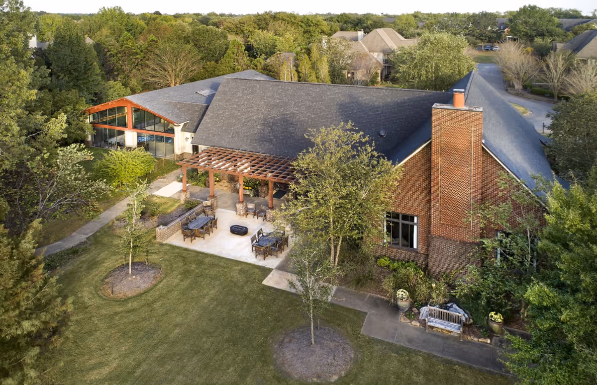 Aerial view of Oak Grove Inn showing a large building with a dark roof and brick chimney, surrounded by trees and greenery. There is an outdoor patio area with tables, chairs, and a pergola. A pathway leads through the landscaped lawn with small trees and shrubs.