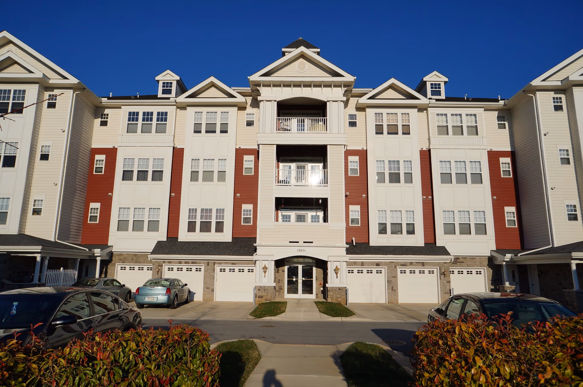Front exterior of a multi-story cream-and-red senior living apartment building with garages, balconies, and parked cars under a clear blue sky.