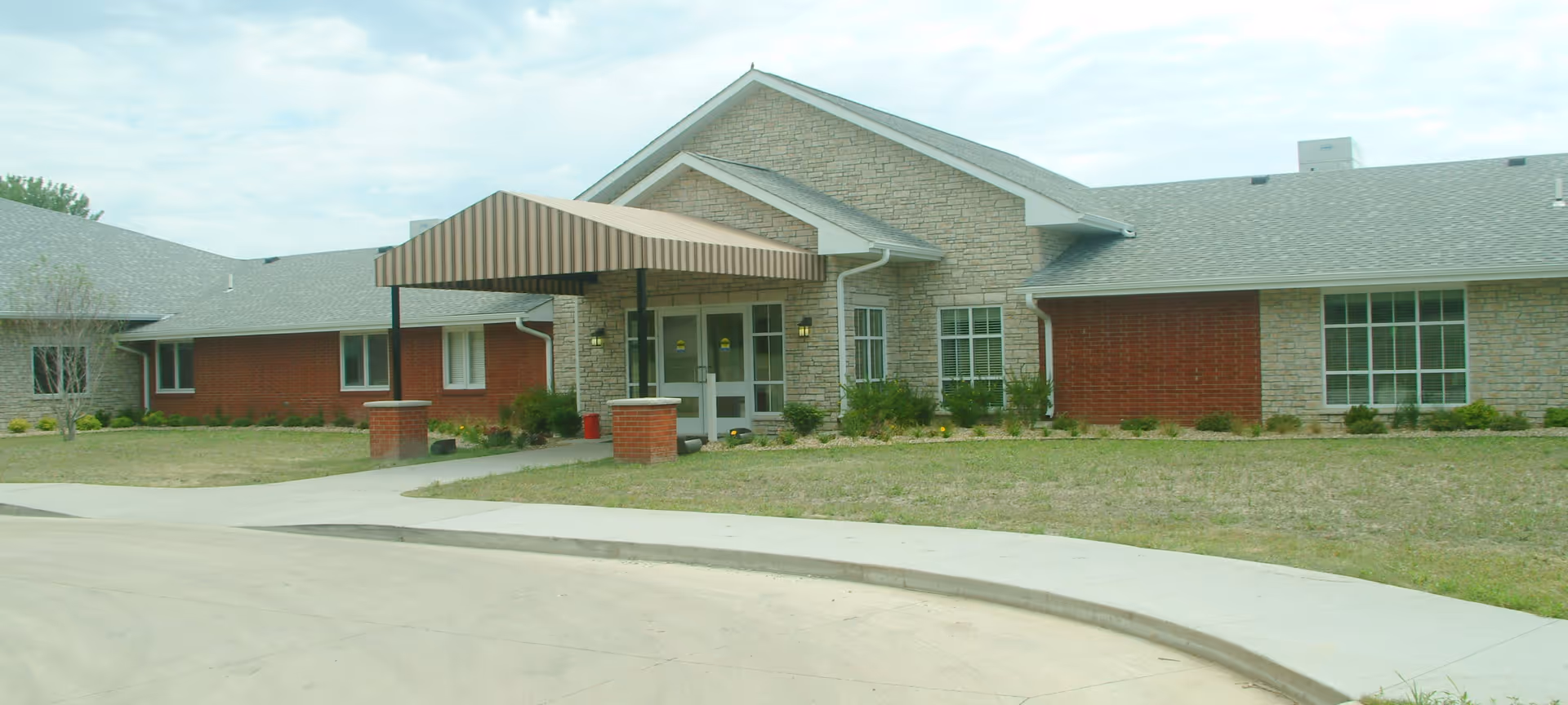 Exterior view of a single-story building with a combination of brick and stone facade. The entrance features a covered awning supported by brick columns. There is a curved driveway and some landscaping with grass and small bushes around the building.