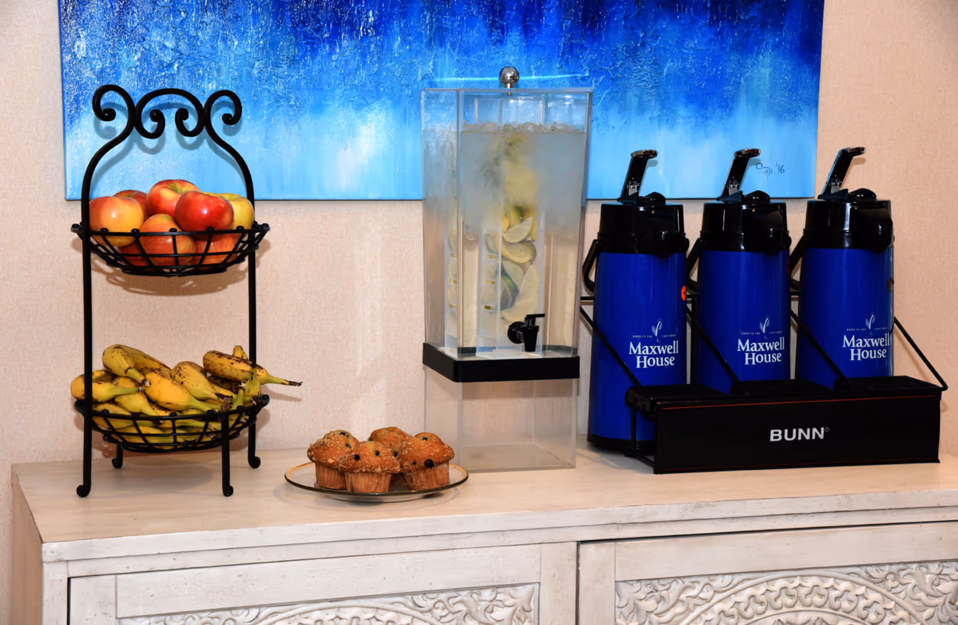 A refreshment station with a two-tiered black metal fruit basket holding apples and bananas, a plate with muffins, a clear beverage dispenser filled with ice and lemon slices, and three blue Maxwell House coffee dispensers on a white decorative cabinet. A blue abstract painting is hanging on the wall behind the setup.