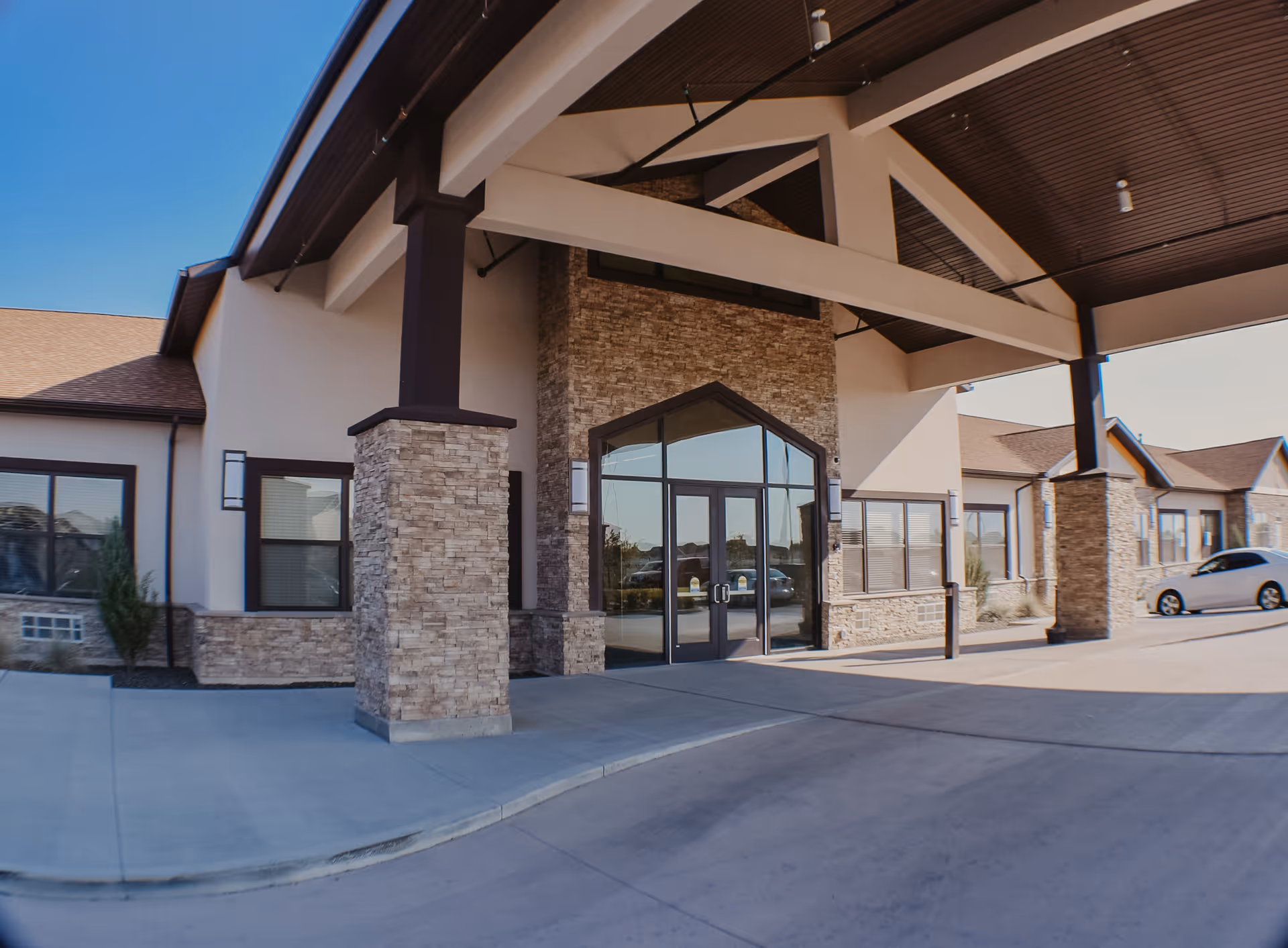 Front exterior view of Serenity Transitional Care facility showing a covered entrance with stone pillars, large glass double doors, and windows on either side under a clear blue sky.