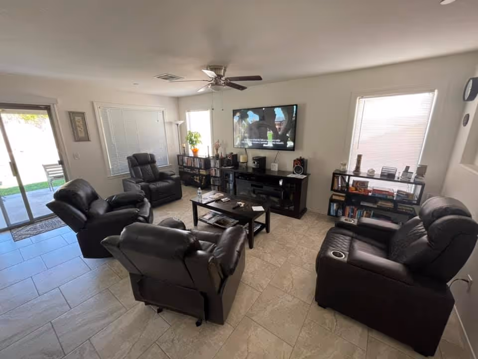 A cozy living room with four black leather recliners arranged around a dark wooden coffee table. A flat-screen TV is mounted on the wall above a media console with speakers and decorative items. Two bookshelves filled with books and decor flank the TV. The room has tiled flooring, two windows with blinds, a ceiling fan, and a sliding glass door leading outside.