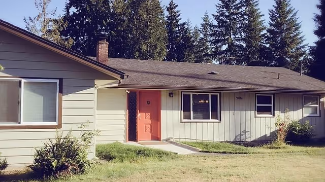 Single-story ranch-style building with a red front door, multiple windows, a grassy lawn, and tall trees in the background.