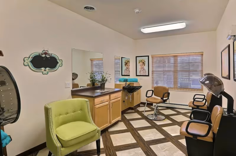 A small salon-style interior with styling chairs, a hooded hair dryer, wash sink, cabinets, mirrors, and a green armchair.