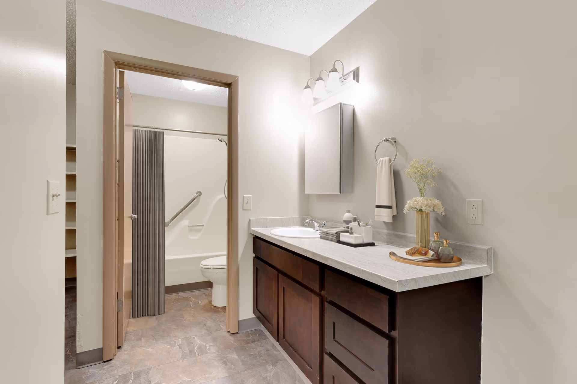 A bathroom with a large vanity featuring a marble countertop, a sink, and a mirror with three light fixtures above it. There is a towel hanging on a ring and a decorative tray with flowers and toiletries on the counter. In the background, there is a doorway leading to a shower and toilet area with a grab bar and a striped shower curtain.