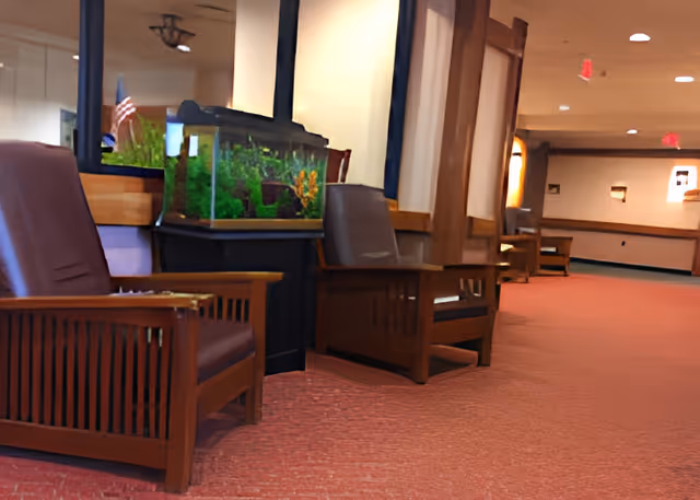 A hallway in a senior living facility with wooden armchairs lined up along the wall. There is a large fish tank on a stand between two chairs. The floor is carpeted in red, and the walls are light-colored with wooden trim. The hallway extends into the distance with more chairs and soft lighting.