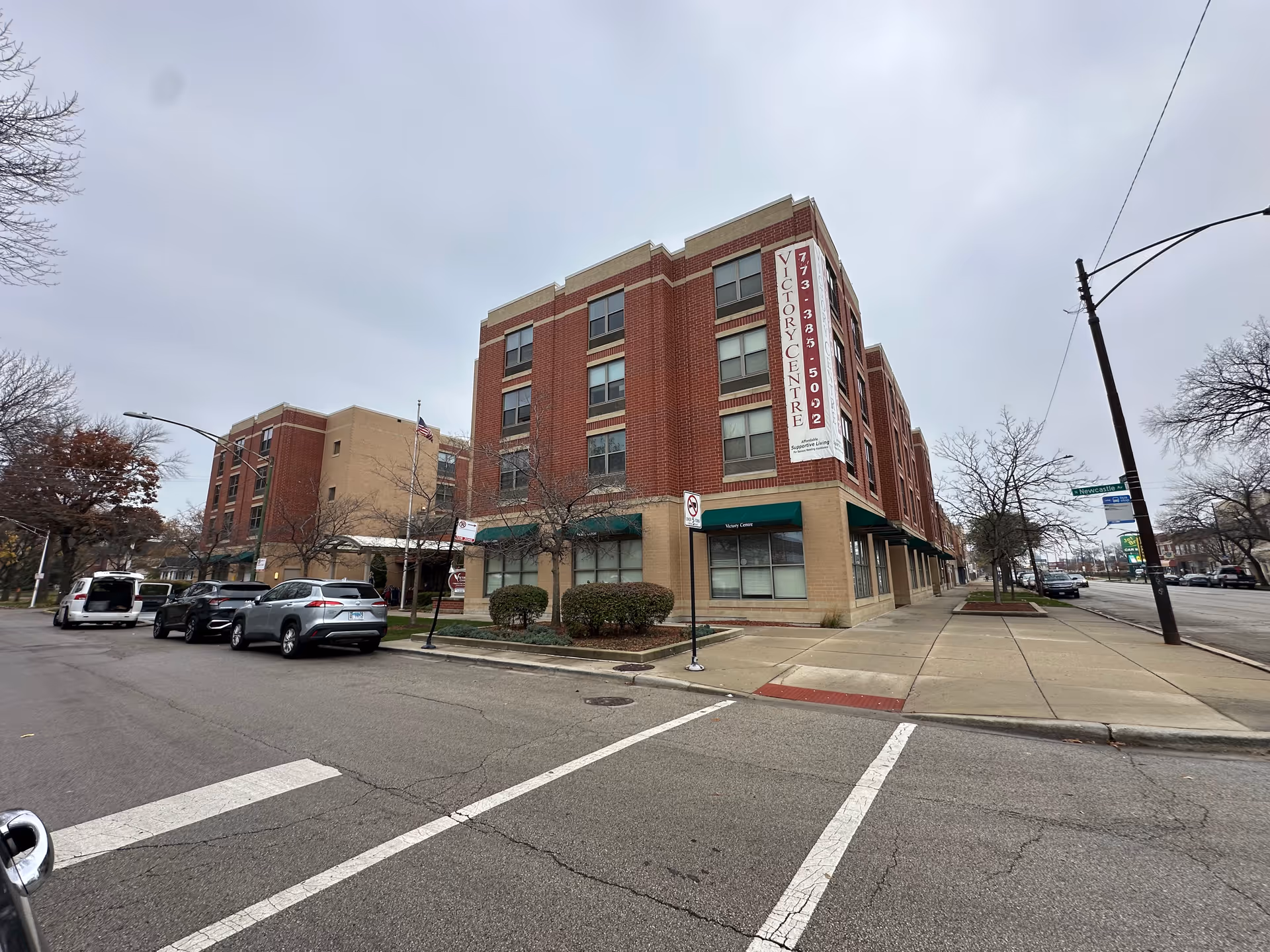 Four-story brick senior living building on a city corner with a vertical 'Victory Centre' banner, parked cars, and a sidewalk under an overcast sky.