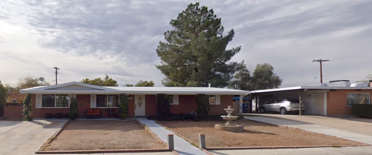 Front exterior view of a single-story brick building with a white roof and a central entrance door. There are two large windows on either side of the door, some greenery climbing the walls, and a small fountain in the front yard. A carport with a silver car is visible on the right side of the building. The sky is partly cloudy.