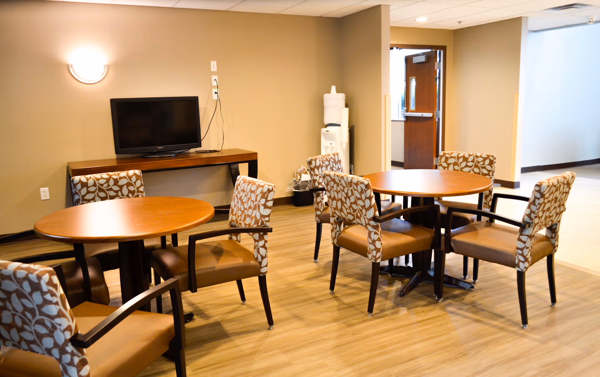 A common area with two round wooden tables surrounded by patterned cushioned chairs. A flat-screen TV is mounted on a wooden console against a beige wall. A water cooler is visible in the corner near an open door leading to another room.
