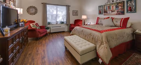 Well-lit furnished bedroom with a large bed, tufted bench at the foot, two red armchairs, a dresser with TV, and a window.