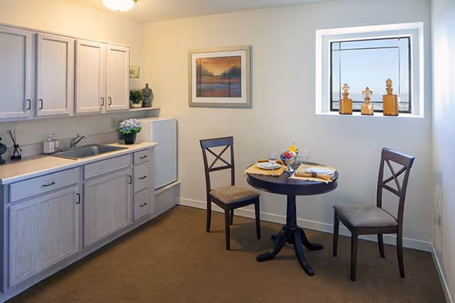 A small kitchen and dining area with light wood cabinets, a sink, and a mini refrigerator. There is a round wooden table set for two with plates, glasses, and a bowl of fruit. Two wooden chairs with cushioned seats are placed around the table. A window with decorative items on the sill and a framed picture on the wall are also visible.