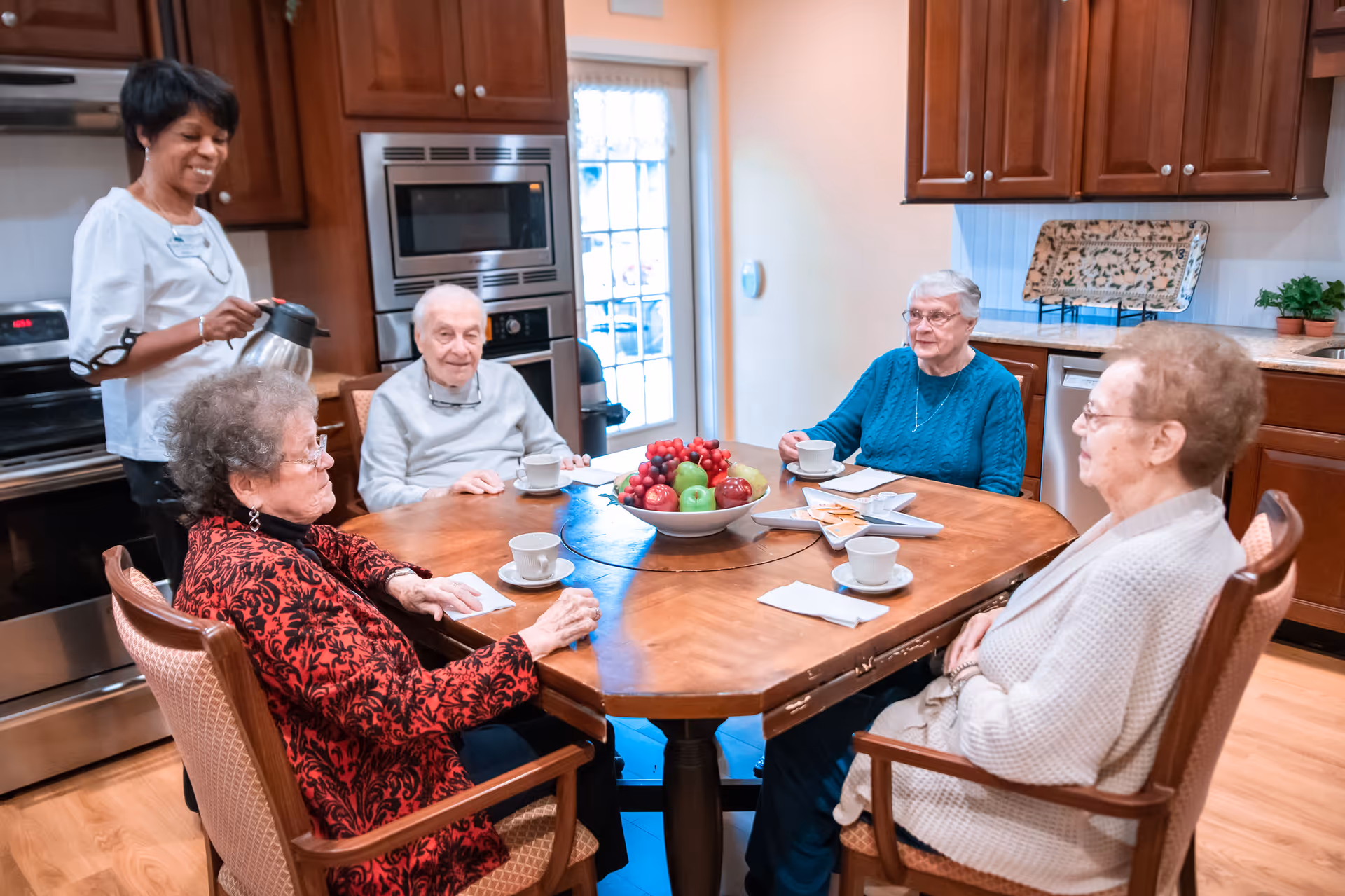 Five older adults seated around a wooden dining table in a kitchen while a staff member pours coffee.