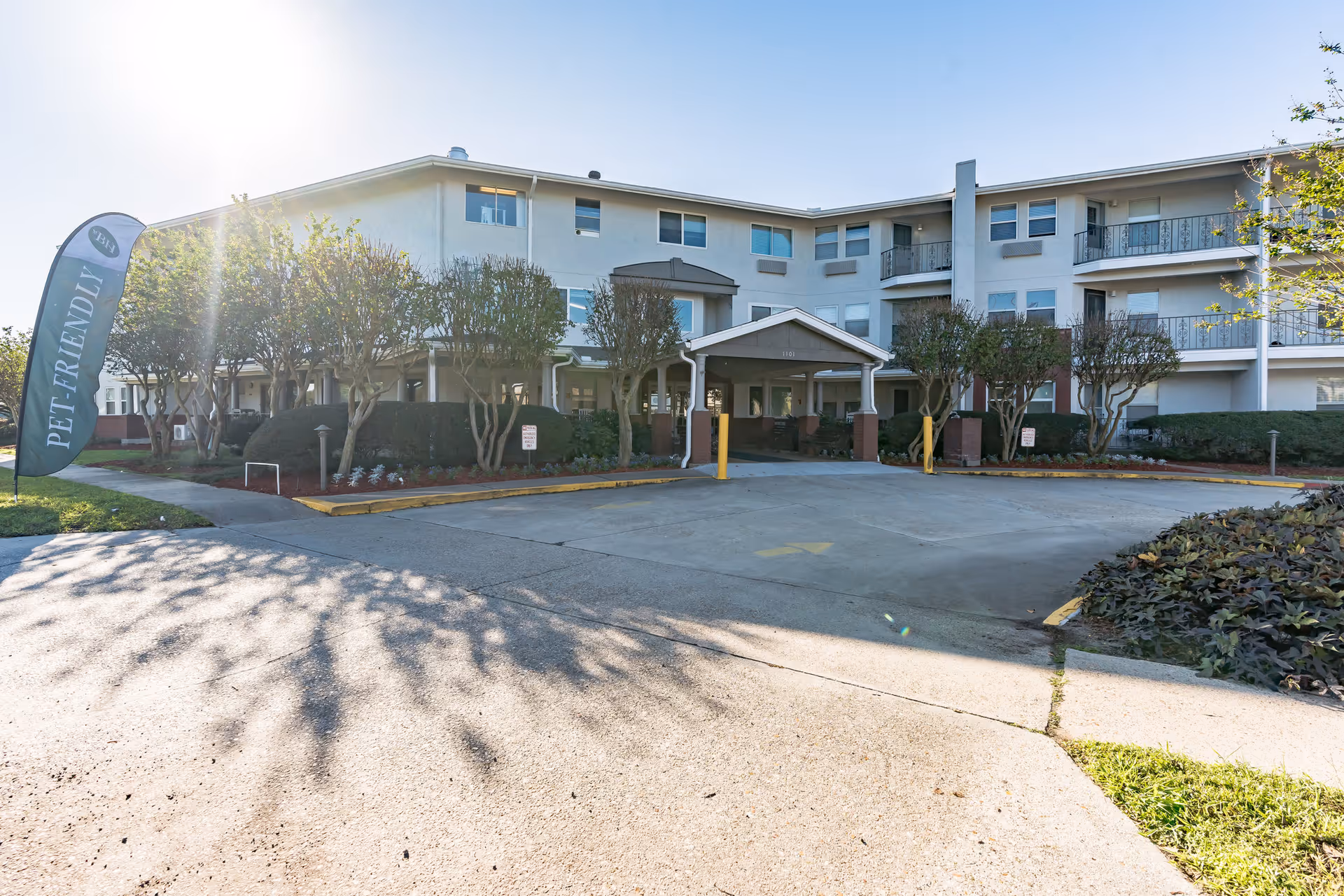 Exterior view of a three-story senior living facility building with balconies and a covered entrance. There are trimmed trees and bushes along the front, a driveway leading to the entrance, and a flag on the left side that reads 'PET-FRIENDLY'. The sun is shining brightly in the sky.